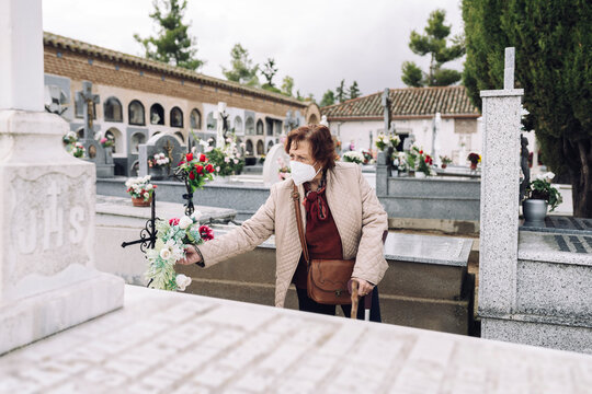 Senior woman wearing face mask putting flowers on grave in cemetery