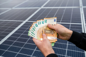 beautiful young hands in black hold a sum of euros in front of solar panels.