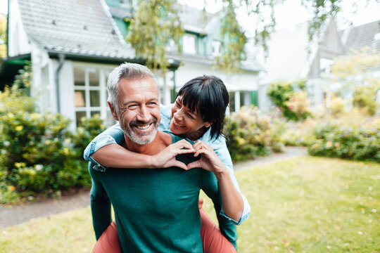 Cheerful man giving piggyback ride to woman gesturing heart shape in backyard - Powered by Adobe