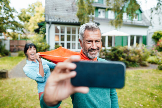 Cheerful woman pulling scarf of man taking selfie on mobile phone