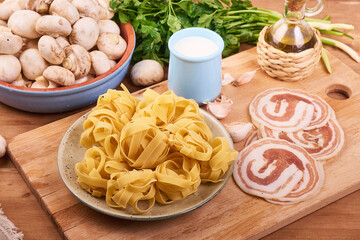 The ingredients for making pasta with creamy mushroom sauce on the kitchen table are fettuccine, mushrooms, cream, olive oil, herbs and pancetta.
