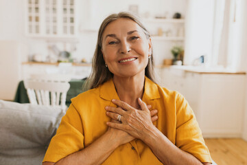 Horizontal indoor image of stylish senior woman in yellow jacket doing grateful gesture, putting...