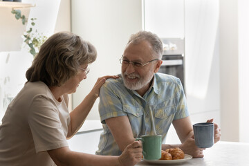 Joyful senior woman talking with laughing old husband, enjoying carefree weekend breakfast time drinking hot coffee or tea eating fresh baked croissants in modern kitchen, communicating concept.