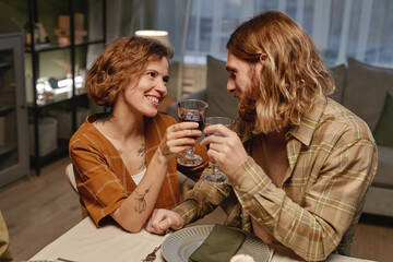 Young happy couple toasting with glasses of red wine while sitting at the table and celebrating the holiday