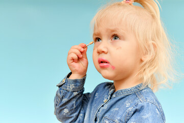 Little blond girl child paints face with eyeshadow. Funny and cheerful children wear makeup for adults.