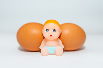 Miniature newborn baby toy and two eggs sitting on a white gray background.Selective focus,copy space.