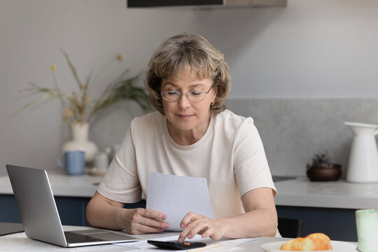 Focused pensive middle aged mature woman analyzing paper bills, calculating domestic utilities or expenditures, involved in financial paperwork, planning investment or paying insurance alone at home.