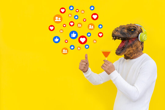 Cheerful surreal man in dinosaur head mask drinking cocktail with social media emoticons isolated on yellow background