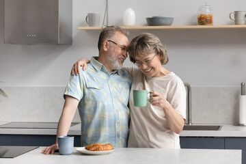 Fototapeta na wymiar Loving affectionate happy middle aged old retired couple cuddling, drinking hot coffee or tea enjoying pleasant conversation standing at table in modern kitchen, good family relations concept.
