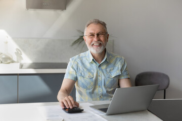 Portrait of happy middle aged 60s man in eyeglasses sitting at table, feeling excited analyzing paper bills, planning budget or making payments online using computer, successful pension investment.