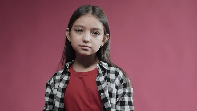 Portrait of little unsatisfied girl looking at camera, an empty face emotion. Cute kid standing in the studio with pink background. High quality 4k footage