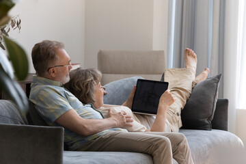 Happy affectionate middle aged senior family couple resting together on cozy sofa, using laptop watching movie or funny photo video online, shopping in internet store, modern tech addiction concept.