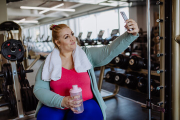 Fototapeta premium Happy mid adult overweight woman taking selfie after exercise indoors in gym
