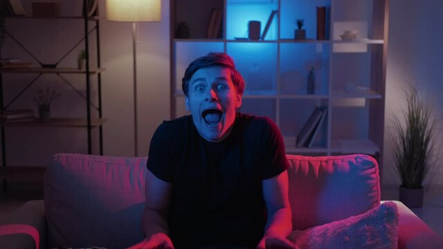 Grimacing man. Meme expression. Silly behavior. Foolish guy making nuts scare face sitting sofa in dark neon light home interior looped.