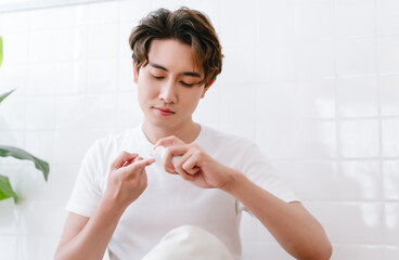 Asian man pumping out cream from white bottle on forefinger after take a bath at bathroom. Young korean or japanese male use moisturizing face balm for healthy glowing skin. Beauty, Body Care Concept.