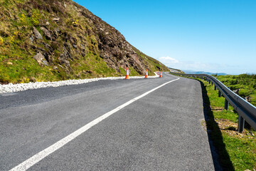 Small narrow asphalt road in a mountains after repair. High quality road surface. Warm sunny day. Metal safety barrier on the right side, hill with grass on a left. Rich blue sky in the background