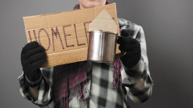 Vagabond woman holds a cardboard sign with the inscription homeless and begging with a steel cup. Male's hand put a cardboard mini-house into mug. Studio shot. The concept of helping vagrants.