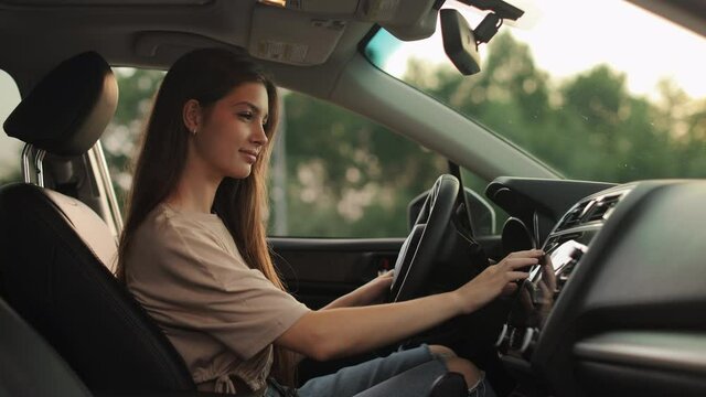 A young woman is sitting in the driver's seat. She is selecting music in the car's multimedia. She is smiling. Shooting from the side seats. 4K 50fps