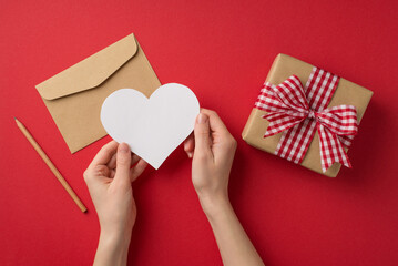 First person top view photo of valentine's day decor girl's hands holding paper heart over envelope pencil and craft paper giftbox with checkered ribbon bow on isolated red background with copyspace