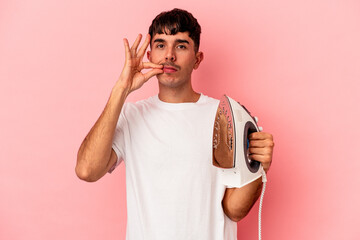 Young mixed race man holding an iron isolated on pink background with fingers on lips keeping a secret.