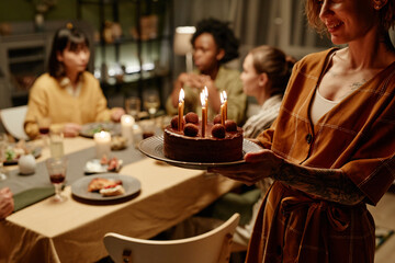 Young woman bringing birthday cake with candles for her friend during birthday party at home
