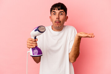 Young mixed race man holding an iron isolated on pink background shrugs shoulders and open eyes...
