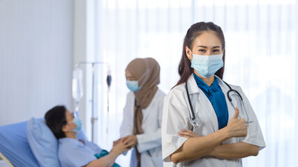 Young asian female doctor wear face mask arms crossed and showing thumbs up while standing in font of muslim doctor checking elderly patient on background. medical team working in the hospital room.