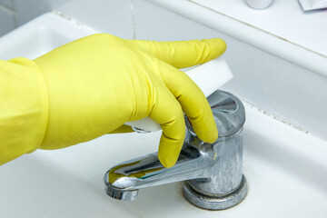 Cleaning the bathroom with melamine sponge. A gloved hand holds a sponge, wipes the plumbing from stains and lime, limescale