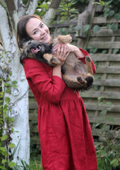 Smiling young woman holds funny border terrier dog. Female in red dress with cute dog. Summer outdoor photo. Playing with puppy in the garden. 