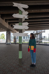 A teenage girl in multicolored trendy clothes stands confusedly near a sign with empty arrows in opposite directions