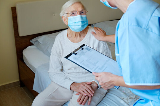 Hospital Nurse Wearing A Face Mask, Holding A Clipboard With Medical History For An Elderly Lady. Senior Woman And Her Designated Care Giver Discussing Test Results. Background, Close Up, Copy Space.