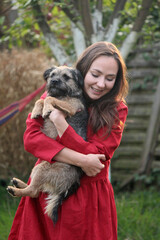 Naklejka na ściany i meble Smiling young woman holds funny border terrier dog. Female in red dress with cute dog. Summer outdoor photo. Playing with puppy in the garden. 