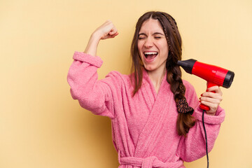 Young woman holding a hairdryer isolated on yellow background raising fist after a victory, winner concept.