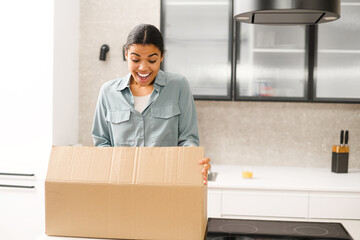 Multiracial woman unpacking parcel cardboard box with online order. Satisfied biracial young woman unpacking parcel box while standing at the kitchen at home. Excited woman looking at delivered goods