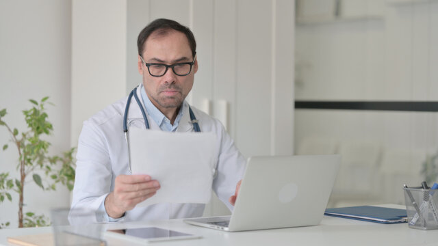 Middle Aged Doctor with Laptop reading Documents in Clinic