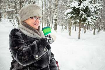 Fototapeta na wymiar An elderly woman drinks coffee while sitting on a bench in a winter park.