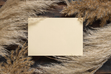 Blank paper card near dried pampas grass on brown wood top view. Boho mockup