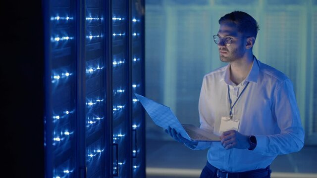 Male Server Engineer in Data Center. IT engineer inspecting a secure server cabinet using modern technology laptop coworking in data center.