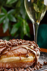 Graffa fritta guarnita con nutella, cioccolato bianco, praline di cioccolato bianche e al latte e con zucchero a velo servita su un piatto nero e con un calice di prosecco