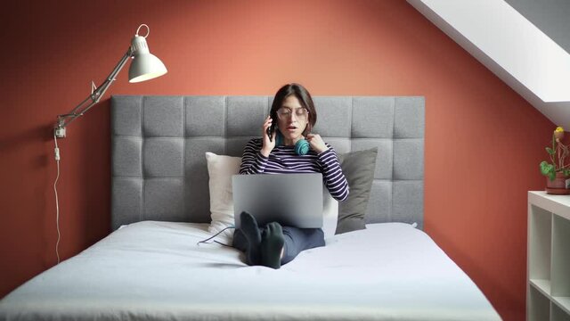 Young female professional working from home. Busy woman sitting on her bed while working with her laptop and taking a phone call in an modern minimal house.