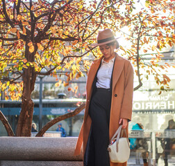 Young asian lady in beige coat and hat calls on mobile phone on city street in park