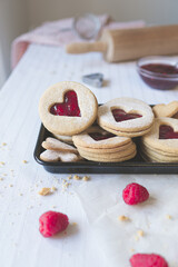 Portrait angled shot of three heart shape linzer cookies and raspberry jam on a white background with baking tools in background. Valentines day, love and romance food concept image.