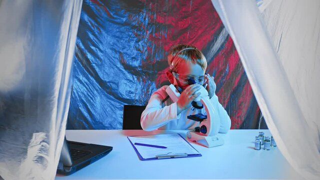 A little boy in a protective mask is playing scientist, looking through a microscope and writing down the results of his observations