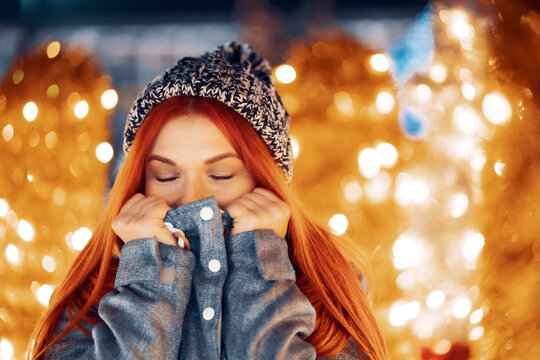 Outdoor night photo of young beautiful happy smiling girl enjoying festive decoration, posing in Christmas fair, in street of european city, wearing knitted beanie hat