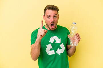 Young caucasian man holding a bottle of plastic to recycle isolated on yellow background having an...