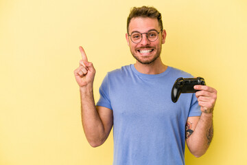 Young caucasian man holding a game controller isolated on yellow background smiling and pointing...