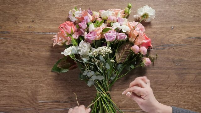 Professional florist woman ties spring bouquet of fresh colorful flowers with thin flexible floral rope on wooden table close upper view.