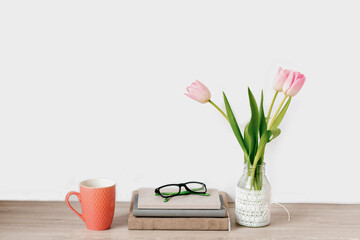 A bouquet of pink tulips of spring flowers in a vase stands on the desktop next to notebooks with glasses and a mug of tea. Copy space