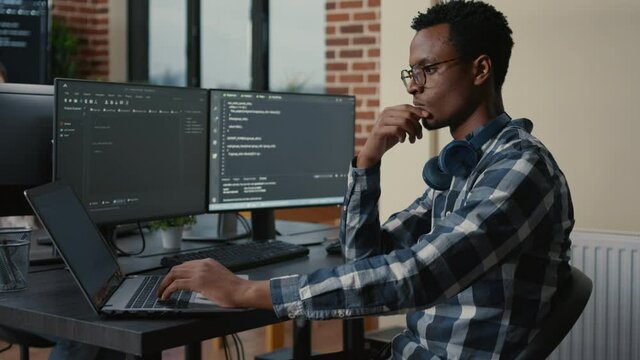 Programer thinking while touching beard and fixing glasses while typing on laptop sitting at desk with multiple screens parsing code. Focused database admin working with team coding in the background.