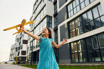 With toy plane. Cheerful little girl walking outdoors in the city in dress